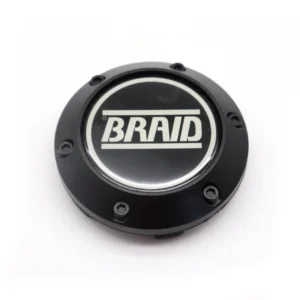 A high-angle studio shot shows a circular, black matte wheel center cap resting on a white surface. The cap features a glossy black center emblem with the word "BRAID" in bold, white, capital letters between two horizontal lines. Six small, black hexagonal bolts are spaced evenly around the outer matte rim, giving the piece a rugged, industrial appearance.